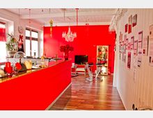 Ein heller, modern gestalteter Innenraum mit auffällig roten Wänden und einer roten Theke. Der Raum enthält dekorative Elemente wie einen Kristallleuchter, Wandbilder und Pflanzen auf der Theke sowie eine kleine Sitzecke mit einem Fernseher.