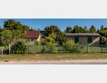 Zwei kleine Wohnhäuser auf benachbarten Grundstücken, eines mit rotem Ziegeldach und das andere mit grün gestrichenen Fensterläden. Die Grundstücke sind von einem grünen Metallzaun umgeben und von Bäumen und Sträuchern bewachsen, im Hintergrund dichter Baumbestand und blauer Himmel.