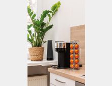 Moderne Küchenzeile mit einer schwarzen Kapsel-Kaffeemaschine auf einer holzfarbenen Arbeitsplatte, daneben ein silberner Ständer mit orangen Kaffee-Kapseln. Im Hintergrund stehen eine grüne Zimmerpflanze in einem geflochtenen Topf und ein schwarzer Lautsprecher auf der Fensterbank.