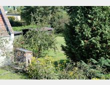 Ein begrünter Garten mit Obstbäumen, großen Nadelbäumen und einer Rasenfläche. Im linken Bildbereich ist ein Haus mit Ziegeldach sowie ein Stapel Feuerholz zu sehen.