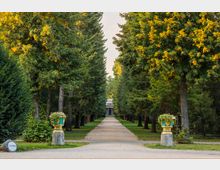 Ein gerader Weg führt durch eine von hohen Bäumen gesäumte Allee zu einem kleinen Gebäude mit Säulen im Hintergrund. Zwei dekorative Pflanzgefäße auf Steinsockeln stehen auf beiden Seiten des Weges, umgeben von gepflegtem Grün.