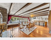 Großer Wohn- und Essbereich mit Holzboden, sichtbaren Deckenbalken und einem Esstisch mit Stühlen auf einem Teppich. An der Seite befinden sich ein gemauerter Kamin sowie eine Bar mit Hockern und Regalen; große Fenster mit roten Vorhängen öffnen den Blick in einen Garten.