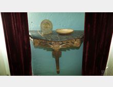 Wandkonsole mit marmorierter Ablage und vergoldeten, geschnitzten Verzierungen, darauf eine goldene Schale und eine runde Metalluhr; die Szene ist von weinroten Vorhängen gerahmt, die Wand ist türkisfarben gestrichen.