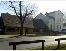 Zwei Gebäude an einer Straße, ein niedrigeres gelbes Haus mit schrägem Ziegeldach und ein höheres, weißes Gebäude im klassischen Stil mit rechteckigen Fenstern; ein kahler Baum steht vorne, und Autos sind im Hintergrund geparkt.