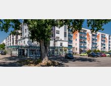 Mehrstöckiges Wohngebäude mit Balkonen und orange-weißen Fassadenelementen, im Vordergrund eine Apotheke mit verglaster Überdachung. Ein Parkplatz mit geparkten Autos und ein großer Baum prägen den Vordergrund der Szenerie.