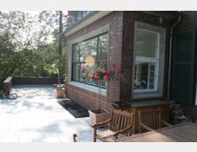 Backsteinfassade eines Gebäudes mit großen Fenstern, davor eine Terrasse mit hellen Steinfliesen. Auf der Terrasse stehen holzfarbene Gartenmöbel, Blumenkübel mit Rosen und im Hintergrund sind Bäume sichtbar.