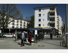 Straßenansicht am U-Bahnhof Bayerischer Platz in Berlin mit einem weißen Wohngebäude, das Balkone und ein Geschäft mit dem Schriftzug „Berliner Bank“ zeigt. Im Vordergrund befinden sich Bäume, parkende Autos und Menschen auf dem Gehweg.