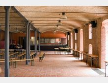 Großer, offener Raum mit Backsteinwänden, einer gewölbten Decke aus Ziegeln und Metallpfeilern. Der Raum ist mit langen Holzbänken und Tischen möbliert, im Hintergrund ist eine Bar zu sehen.