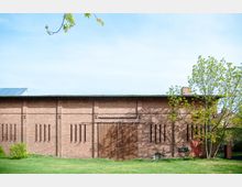 Backsteinbau mit einer großen Holzschiebetür und schmalen, senkrechten Fensteröffnungen, umgeben von einer grasbewachsenen Fläche und einzelnen Bäumen. Das Flachdach ist teilweise mit Solarpaneelen bedeckt.