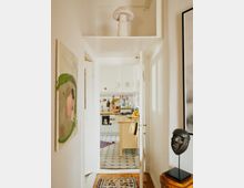 Ein schmaler, heller Flur mit einem Läufer führt zu einer offenen Küche mit weiß-blauen Bodenfliesen. Ein hölzerner Küchentisch steht in der Mitte der Küche; Dekorationen wie ein Holzstuhl mit Hut, eine afrikanische Maske auf einem Podest und Bilder an den Wänden prägen den Raum.