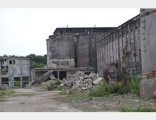 Verlassene Industrieanlage mit zerfallenen Gebäuden, großen Betonsilos und Schutthaufen. Im Vordergrund befinden sich überwucherte Trümmer und verbogene Stahlträger, während im Hintergrund Wälder sichtbar sind.