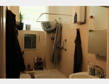 Kleines Badezimmer mit hellbeigen Fliesen, Badewanne mit Duschvorhang und Haltegriff sowie Waschbecken mit Spiegel und Ablage. Ein Fenster mit Pflanzen bringt Tageslicht in den Raum; Handtücher und Badutensilien sind an Haken und Regalen verstaut.