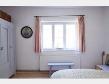 Schlafzimmer mit schlichter Einrichtung, darunter ein weißer Wandschrank, ein Fenster mit gestreiften Vorhängen, eine Heizung darunter und eine blaue Sitzbank. An der Wand hängt ein rundes Gemälde, und auf der Fensterbank steht ein kleines Modell eines Segelboots.
