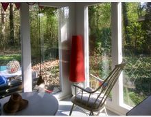 Ein heller Wohnraum mit großen Fenstern, die den Blick auf einen dicht bewachsenen Waldgarten ermöglichen. Im Vordergrund stehen ein weißer Tisch, ein Schaukelstuhl mit Kissen und eine rote Stehlampe, während im Außenbereich ein Tisch mit Dekoration sichtbar ist.