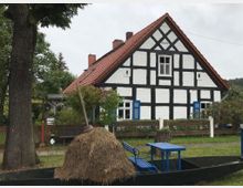 Fachwerkhaus mit weiß verputzten Wänden und schwarzen Balken, roten Dachziegeln und blauen Fensterläden, umgeben von einem Garten mit Bäumen. Im Vordergrund Heuhaufen in einem schwarzen Holzkahn, daneben blaue Sitzmöbel auf einer Wiese.