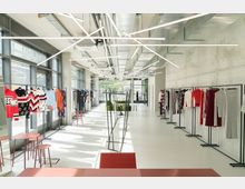 Großer, moderner Innenraum mit Kleidungsstücken auf Kleiderständern entlang beider Seitenwände. Der Raum besitzt eine Betonwand, große Fensterfronten und auffällige, geometrische LED-Leuchten an der Decke.