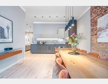 Offener Wohn- und Essbereich mit moderner grauer Einbauküche, langem Holzesstisch und mehreren Polsterstühlen unter schwarzen Pendelleuchten. Helle Holzböden, eingelassene Deckenleuchten, eine freiliegende Ziegelwand und gerahmte Wandbilder prägen den Raum.