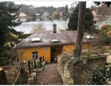 Ehemaliges gelbes Gebäude mit Flachdach, umgeben von Bäumen und einem gepflasterten Weg, der hinunter zum Eingang führt. Im Hintergrund ein See, umgeben von Häusern und bewaldeten Ufern.