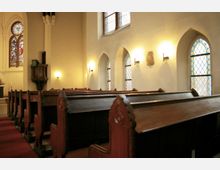 Innenraum einer Kirche mit Holzbänken, einem roten Teppichboden und farbigen Glasfenstern. Links neben den Bänken sind eine geschnitzte Kanzel und der Altar sichtbar, Wandleuchten beleuchten den Raum.