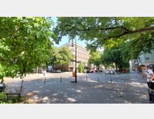 Straßenszene mit gepflastertem Platz, umgeben von Bäumen und einem historischen Gebäude mit heller Fassade im Hintergrund. Auf dem Platz befinden sich Fahrradbügel, Mülleimer und parkende Autos; Passanten sind am Rand des Bildes sichtbar.