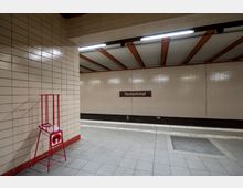 U-Bahn-Station mit hellen, cremefarben gefliesten Wänden und einer Decke mit orangefarbenen Streben. An der Wand befindet sich ein Schild mit der Aufschrift „Nordbahnhof“, und im Vordergrund steht ein roter Ständer mit Informationsmaterial.