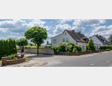 Straßenszene in einem Wohngebiet mit freistehenden Häusern, Zäunen und gepflegten Vorgärten. Ein Gehweg, eine kleine Grünfläche mit Bäumen sowie eine niedrige Mauer sind sichtbar, unter einem bewölkten Himmel.