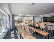 Ein heller Seminarraum mit großen Fensterfronten, durch die Bäume und ein Gebäude im Außenbereich sichtbar sind. Aufgestellte Stühle stehen auf Tischen, ein Whiteboard und ein großer Bildschirm sind an der Wand angebracht.