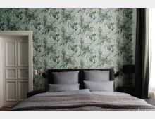 Schlafzimmer mit einer Tapete im grünen Pflanzenmuster, einem grauen Polsterbett mit grauer Bettwäsche und einer grauen Tagesdecke. Links befindet sich eine weiße Tür und rechts eine schwarze Nachttischlampe vor einem halb geöffneten Fenster mit Vorhang.