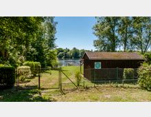 Ein eingezäunter Bereich mit einem Holzhäuschen rechts im Bild, das von Bäumen und Sträuchern umgeben ist. Im Hintergrund ist ein See mit einem Steg und dichter Bewaldung zu sehen, bei sonnigem Wetter.