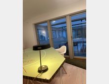 Ein moderner Innenraum mit einem großen Tisch im Vordergrund, der eine grüne Oberfläche mit roten Markierungen hat, beleuchtet von einer schwarzen Schreibtischlampe. Im Hintergrund sind bodentiefe Fenster mit Blick auf einen verglasten Wintergarten oder Balkon zu sehen.