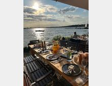 Großer, gedeckter Holztisch im Freien mit eleganter Tischdekoration, darunter Blumen, Stoffservietten und Glasflaschen, am Ufer eines Sees mit Blick auf das Wasser, unter einem sonnigen Himmel mit Wolken.