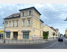 Eine Straßenecke in einer Stadt mit einem beigefarbenen, zweistöckigen Gebäude im Vordergrund, das Schilder von Versicherungs- und Geschäftsfilialen trägt. Die Straße führt entlang einer Reihe von Wohnhäusern mit hellgelben Fassaden und roten Dächern; Laternen und ein geparktes Auto sind sichtbar.