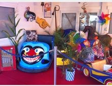 Bunte Ausstellung mit nostalgischen Kirmes-Objekten, darunter ein blaues Autoskooter-Fahrzeug mit clownartigem Gesicht, Holzkarussellpferde, dekorative Pflanzen und andere thematische Dekorationen, auf rotem Teppich präsentiert. Im Hintergrund sind Fenster, ein Plakat und eine hängende Handattrappe zu sehen.