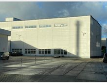 Großer grauer Industriegebäude-Komplex mit mehreren Fenstern in der oberen und unteren Etage. Davor ein gepflasterter Hof mit vereinzelten Autos und Absperrpfosten, von der Sonne und dem Schatten eines Schornsteins beleuchtet.