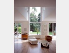 Helles Wohnzimmer mit hohen Decken und großen bodentiefen Fenstern, die auf einen grünen Garten führen. Der Raum ist mit einem Sessel, einem Couchtisch aus Glas und einem Holzboden ausgestattet; Regale und ein Sideboard ergänzen die minimalistische Einrichtung.