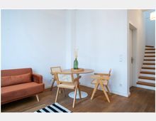 Minimalistisch eingerichtetes Wohnzimmer mit Holzboden, einem orangefarbenen Sofa, einem runden Holztisch mit drei Holzstühlen und einer grünen Vase als Dekoration. Rechts führt eine Treppe mit Holzstufen nach oben, und eine Tür ist neben der Treppe erkennbar.