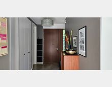 Eingangsbereich eines modernen Hauses mit grauen Wänden, einer Holzeingangstür und Einbauschränken auf der linken Seite. Ein schmaler Holzschrank auf der rechten Seite ist mit einer Vase mit Blumen und dekorativen Schalen versehen; darüber hängen gerahmte Bilder.