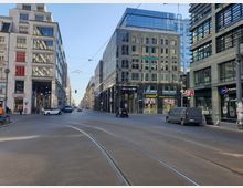 Straßenszene in einer urbanen Umgebung mit modernen und historischen Gebäuden. Im Vordergrund verlaufen Straßenbahnschienen, während Autos und Fußgänger sichtbar sind; Geschäfte und Schaufenster befinden sich entlang der Gebäude auf beiden Seiten.