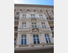 Fassade eines historischen Wohngebäudes mit reich verzierten Stuckelementen, weißen Fensterrahmen und breiten Balkonen mit kunstvoll gestalteten Metallgeländern, vor einem blauen Himmel.