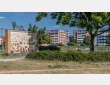 Eine städtische Grünfläche mit einem Baum im Vordergrund und einem Mosaikbild aus Keramik auf einer Backsteinwand. Im Hintergrund stehen mehrstöckige Wohnhäuser mit Balkonen, umgeben von weiteren Bäumen und Rasenflächen.