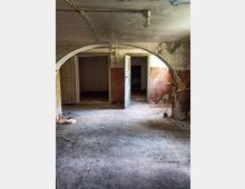 Ein leerer, heruntergekommener Innenraum mit sichtbaren Schäden an den Wänden und der Decke, darunter Risse und abblätternder Putz. Im Vordergrund ist ein schmutziger Boden, im Hintergrund mehrere geöffnete Türen, die in weitere verlassene Räume führen.