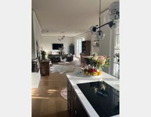 Wohnbereich mit offener Küche in modernem Stil. Zu sehen sind eine Kücheninsel mit weißer Arbeitsplatte und Dekorationen, ein Esstisch mit Stühlen, ein Sofa und ein Fernseher im hinteren Bereich, sowie große Fenster mit viel Tageslicht.