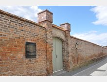 Ziegelmauer mit einem Torbogen und geschlossener hölzerner Doppeltür, flankiert von zwei Backsteinsäulen. Links an der Wand befindet sich eine Gedenktafel, und rechts ist eine historische Laterne zu sehen.