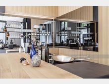 Moderne Küche mit Holz- und schwarzen Hochglanzflächen, zentraler Kücheninsel mit Dekorationsobjekten und Küchenutensilien auf einer Holzarbeitsplatte. Im Hintergrund offene Regale mit Gläsern und Tassen sowie Küchengeräte und ein Spülbecken mit Armatur.