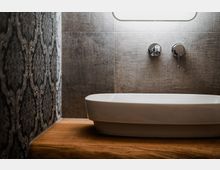 Modernes Badezimmer-Waschbecken aus weißer Keramik auf einer Holzablage, davor eine Wand aus grauen Fliesen mit wandmontiertem Wasserhahn und Bedienhebel. Links ist eine dunkle, gemusterte Tapete als Kontrast zur Fliesenwand zu sehen.