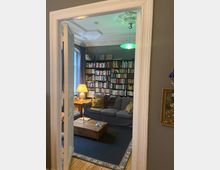 Blick durch eine Tür in ein Wohnzimmer mit hoher Stuckdecke, grünem Hängelicht und einer wandfüllenden Bücherregalwand. Im Raum stehen ein graues Sofa, ein niedriger Holztisch auf einem großen Teppich sowie ein Beistelltisch mit Lampe neben einem Fenster.