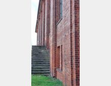 Ziegelsteinfassade eines Gebäudes mit vertikalen Stützelementen, daneben eine schmale, seitlich verlaufende Steintreppe; im Vordergrund eine kleine Grasfläche.