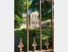 Historisches Gebäude aus Sandstein mit blauen Fensterläden, umgeben von einem gepflegten Garten mit blühenden Sträuchern und großen Bäumen, gesehen durch ein verziertes, schmiedeeisernes Tor.