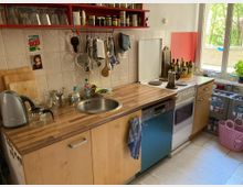 Kleine Küche mit Holzarbeitsplatte, Spüle, Herd und Hängeregalen. Auf der Arbeitsfläche stehen diverse Küchenutensilien, Geschirr und Flaschen, während ein großes Fenster den Raum mit Tageslicht erhellt.