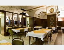 Restaurant mit klassischem Interieur, hell erleuchtet. Holzwände, dekorative Deckenverzierungen, weiße Tischdecken mit gelben Tischläufern und braune Stühle prägen das Ambiente.